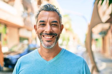 Orta yaşlı, kır saçlı, gülümseyen bir adam şehrin caddesinde mutlu bir şekilde yürüyor..