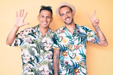 Genç eşcinsel çift, yaz şapkası ve Hawaii tişörtü takmış yedi numaralı parmaklarıyla gösteriş yapıyor ve gülümsüyor.. 