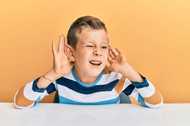 Kafkasyalı sevimli bir çocuk, günlük giysiler içinde masada oturmuş iki elini de kulak hareketi yaparken duymaya çalışıyor, dedikoduya meraklı. İşitme sorunu, sağır 