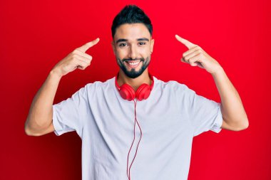 Sakallı genç adam kulaklıkla müzik dinliyor iki eliyle başını gösteren gülümsemeler, harika bir fikir ya da düşünce, iyi bir hafıza 