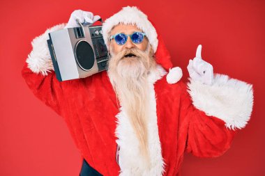 Old senior man wearing santa claus costume and boombox surprised with an idea or question pointing finger with happy face, number one 