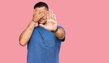 Dövmeleri olan, sıradan kıyafetler giyen, elleriyle gözleri kapatan, üzgün ve korku ifadeli hareketler yapan yakışıklı bir adam. utangaç ve olumsuz kavram. 