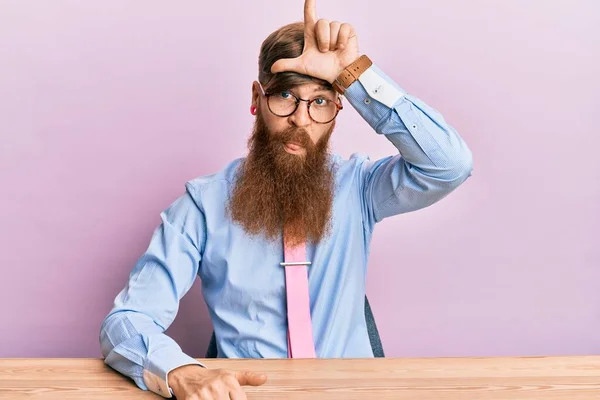 Young irish redhead man wearing business shirt and tie sitting on the table making fun of people with fingers on forehead doing loser gesture mocking and insulting.