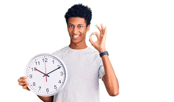 Młody Afrykanin Amerykański Człowiek Trzyma Duży Zegar Robi Znak Palcami — Zdjęcie stockowe