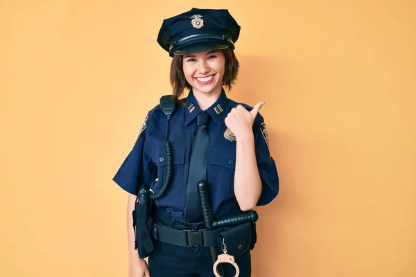Young beautiful woman wearing police uniform smiling with happy face looking and pointing to the side with thumb up.