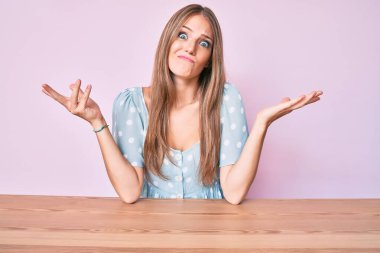 Gündelik kıyafetler giyen genç sarışın kız masanın üzerinde elleri ve kolları havada, kafası karışmış bir şekilde oturuyor. şüphe kavramı. 