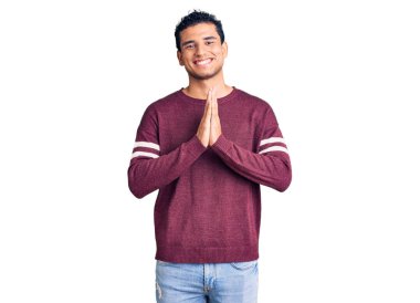 Hispanik yakışıklı genç adam gündelik giysiler içinde ellerini birleştirip dua ediyor af diliyor kendinden emin bir şekilde gülümsüyor.. 