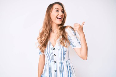 Sarışın, yaz elbisesi giymiş, beyaz, genç bir kadın baş parmağını yana kaldırmış, ağzı açık, mutlu bir şekilde gülümsüyor. 