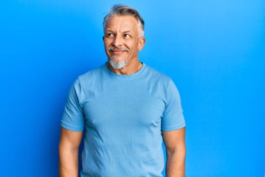 Orta yaşlı, gri saçlı, günlük kıyafetler giyen, gülümseyen, yan bakan ve düşünen bir adam.. 