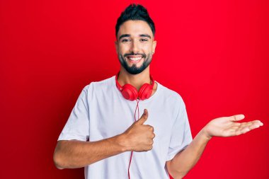 Sakallı genç adam kulaklıkla müzik dinliyor, avuç içi gösteriyor ve baş parmaklarıyla iyi bir hareket yapıyor, mutlu ve neşeli gülümsüyor. 