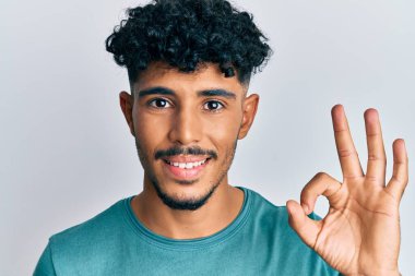 Gündelik giysiler giyen genç Arap yakışıklı adam parmaklarıyla imza atıyor, gülümseyerek mükemmel bir sembol oluşturuyor. 