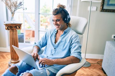 Evde dizüstü bilgisayar ve kulaklık kullanarak mutlu gülümseyen genç İspanyol adam.