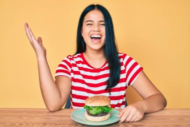 Lezzetli bir klasik burger yiyen Asyalı genç kız zaferi mutlu bir gülümsemeyle ve kazanan yüz ifadesiyle kutluyor. 