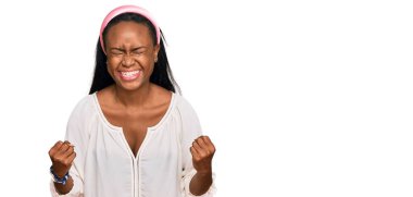 Genç siyah kadın gündelik kıyafetler giyip çok mutlu ve heyecanlı bir şekilde kollarını kaldırıp, gülümseyerek ve başarı için bağırarak kazanan jestler yapıyor. kutlama konsepti. 