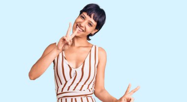 Kısa saçlı, yaz kıyafeti giyen, gülümseyen, parmaklarını zafer işareti yaparken gösteren genç esmer kadın. İki numara.. 