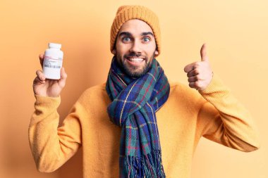 Sakallı, elinde hap şişesi olan, mutlu ve pozitif gülümseyen, başparmağı mükemmel ve onay işareti yapan genç yakışıklı bir adam. 