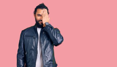 Genç Arap adam deri ceket giyiyor. Bir gözünü eliyle kapatıyor. Yüzünde kendine güvenen bir gülümseme ve sürpriz bir duygu var.. 