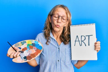 Orta yaşlı sarışın kadın boyacı paleti ve not defteri tutuyor. Kameraya bakıyor. Öpüşme sahnesini çekiyor. aşk ifadesi. 