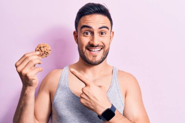 Sakallı, kolsuz tişörtlü, çikolatalı kurabiye yiyen, mutlu, eli ve parmağı olan genç bir adam.