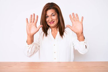 Orta yaşlı esmer, İspanyol iş kadını, gündelik beyaz gömlekli, masanın üzerinde oturuyor ve parmaklarını 10 numarayı göstererek gülümsüyor ve mutlu.. 