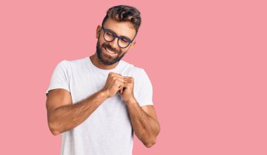 Gündelik giysiler ve gözlük takan genç İspanyol bir adam. Gergin ve heyecanlı bir şekilde gülümsüyor. Elleri çenesine bakıyor. 