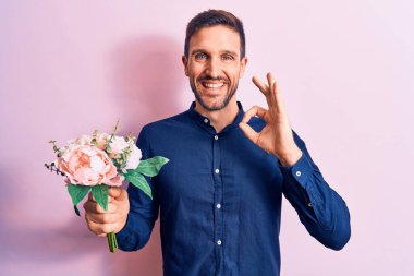 Genç yakışıklı romantik adam pembe arka planda güzel bir buket çiçek tutuyor tamam işareti yapıyor parmaklarıyla, gülümseyerek mükemmel bir sembol oluşturuyor