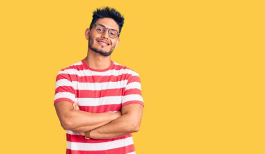 Yakışıklı Latin Amerikalı, sıradan kıyafetler ve gözlük takan mutlu yüzlü, kollarını kavuşturup kameraya bakan genç adam. pozitif kişi. 