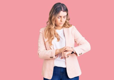 İş kıyafetleri giymiş genç beyaz bir kadın kol saatini kontrol ediyor, rahat ve kendinden emin. 