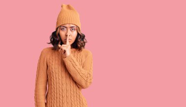 Genç, güzel, melez bir kadın yün kazak ve kışlık şapka giyip dudaklarında parmaklarıyla sessiz olmak istiyor. sessizlik ve gizli konsept. 