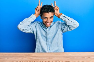 Genç, yakışıklı, Afro-Amerikan bir adam, gündelik giysiler içinde masada oturmuş parmakları tavşan kulaklı, komik ve çılgın poz veriyor, neşeli gülümsüyor. 