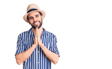Sakallı, yaz şapkalı ve çizgili tişörtlü genç bir adam ellerle dua ederek af diliyor kendinden emin bir şekilde gülümseyerek.. 