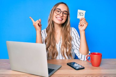 Genç, sarışın bir kadın elinde evet hatırlatıcısı masaya oturmuş gülümsüyor mutlu bir şekilde el ve parmak kenarı işaret ediyor. 