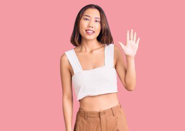 Genç, güzel Çinli kız, günlük kıyafetler giyip beş numarayı gösterip işaret ederken kendinden emin ve mutlu bir şekilde gülümsüyor.. 