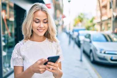 Beyaz kız şehrin caddesinde akıllı telefon kullanarak mutlu bir şekilde gülümsüyor..