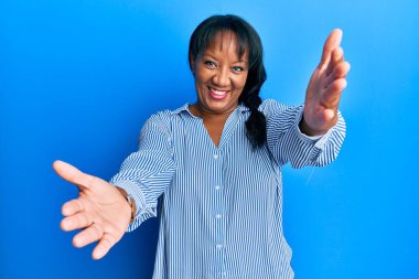 Orta yaşlı Afro-Amerikan kadın günlük giysiler içinde kameraya bakıp kucaklaşmak için gülümsüyor. Mutluluğu kucaklayan neşeli bir ifade.. 