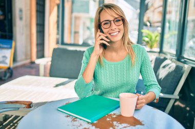 Genç sarışın kadın akıllı telefonda konuşuyor ve kahve dükkanının terasında kahve içiyor..