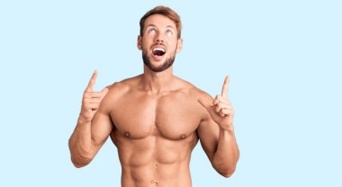 Kafkasyalı genç adam tişörtsüz duruyor. Şaşırmış ve şaşırmış bir şekilde yukarı bakıyor, parmaklarıyla işaret ediyor ve kollarını kaldırıyor.. 