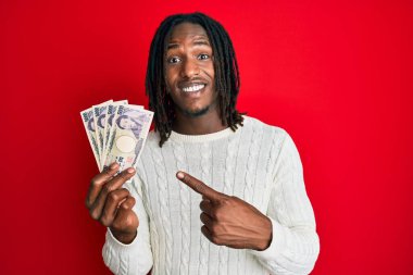 Saçları örgülü Afro-Amerikan bir adam 5000 Japon yenini elinde tutarken mutlu mesut elini ve parmağını göstererek gülümsedi. 