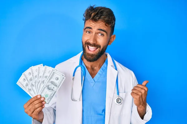 Joven Hispano Vistiendo Uniforme Médico Sosteniendo Dólares Apuntando Con Pulgar — Foto de Stock