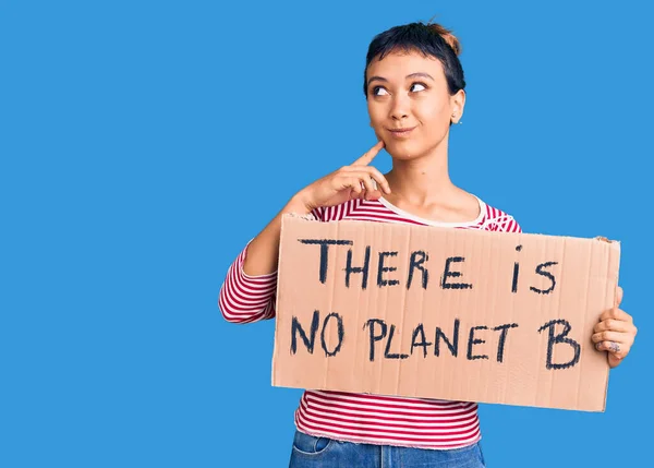 Young woman holding there is no planet b banner serious face thinking about question with hand on chin, thoughtful about confusing idea