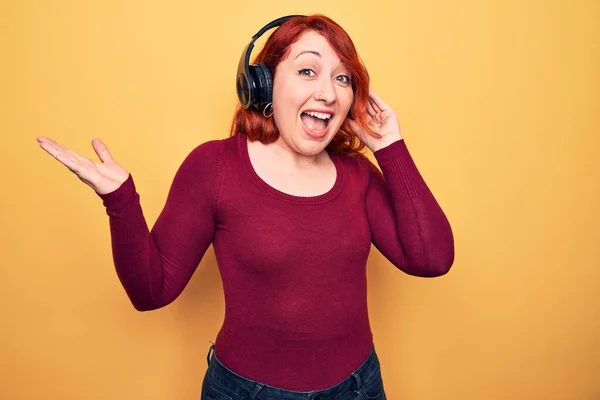 幸せな笑顔で達成を祝う黄色の背景にヘッドフォンを使用して音楽を聴く若い美しい赤毛の女性と手を上げて受賞式 — ストック写真