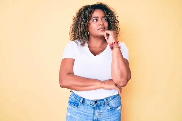 若いアフリカ系アメリカ人の女性には 質問について考える顎に手でカジュアルな白いTシャツを着て 集中的な表現 思慮深い顔で笑顔 疑わしい概念 — ストック写真