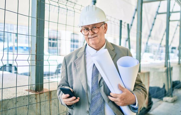 Senior grey-haired architect man holding blueprints using smartphone at street of city