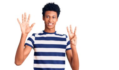 Genç, Afro-Amerikalı, günlük giysiler giymiş, 8 numaralı parmaklarıyla işaret ederken, kendinden emin ve mutlu bir şekilde gülümsüyor.. 