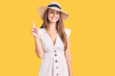 Genç, sarışın, yaz elbisesi ve şapkası giyen güzel bir kadın bir numaralı parmağını gösterip işaret ederken kendine güvenen ve mutlu bir şekilde gülümsüyor.. 