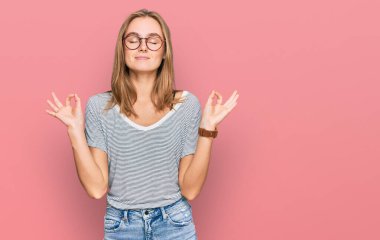 Güzel, genç, sarışın bir kadın günlük kıyafetler ve gözlük takıyor rahatlıyor ve gözleri kapalı gülümsüyor parmaklarıyla meditasyon hareketi yapıyor. yoga konsepti. 