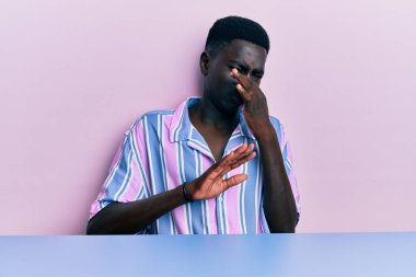 Genç, Afro-Amerikan bir adam, gündelik giysiler içinde masada oturmuş pis ve iğrenç bir koku alıyor. Dayanılmaz bir koku. Nefesini burnunda tutuyor. Kötü kokuyor. 