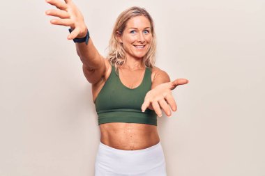 Orta yaşlı beyaz kadın spor giysisi giymiş kameraya bakıp sarılmak için kollarını açmış gülümsüyor. Mutluluğu kucaklayan neşeli bir ifade.. 