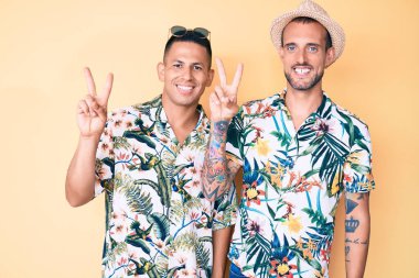 Yazlık şapkalı ve Hawaii tişörtlü iki genç eşcinsel kameraya bakıp zafer işareti yapıyor. İki numara.. 