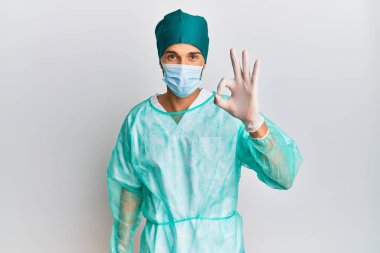 Cerrah üniforması ve tıbbi maske takan yakışıklı genç adam gülümsüyor. El ve parmaklarıyla 